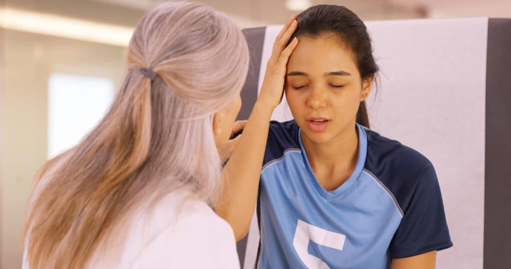 A girl wearing a blue jersey holds her head with one hand. Her eyes are closed and a woman in a white lab coat stands in front of her.