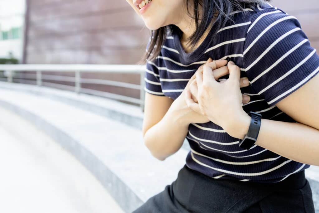 A woman sits down while clutching her left chest with both hands. She appears to be in pain.
