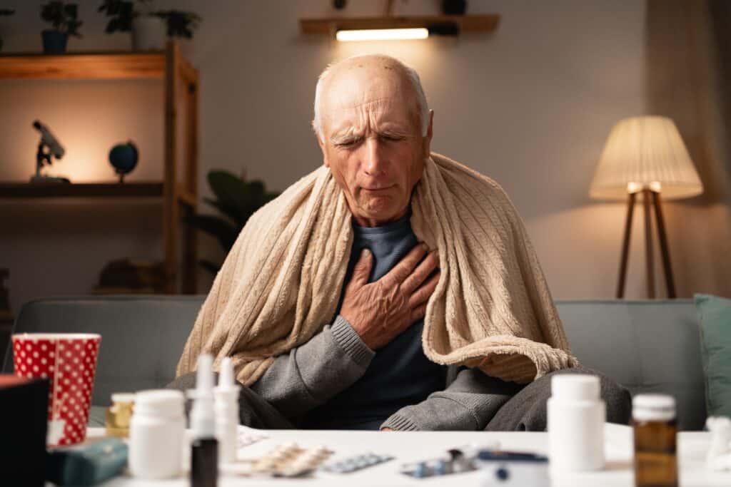 A man sits on a couch with a blanket wrapped around his shoulders. His eyes are closed, and he holds his hand to his chest.