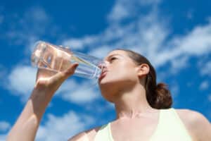 Woman tilting head back while drinking water from a clear bottle on a sunny day